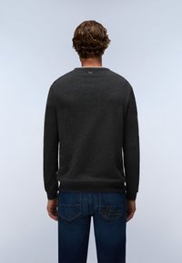 Man with curly hair wearing a dark gray long-sleeve sweater and blue jeans, standing facing away against a plain light background.