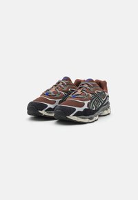Chaussures de sport marron et noires avec des superpositions en maille et en suede, dotées d'une semelle incurvée et d'accents argentés contrastés. Lacets dans une nuance de gris.