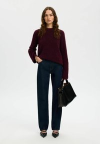 Una donna con lunghi capelli mossi indossa un maglione viola scuro, jeans blu scuro, scarpe nere a punta e porta una grande borsa nera.