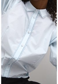 Light blue button-up shirt made of smooth fabric featuring a scalloped trim, round buttons, and a classic collar. Straight fit with long sleeves.