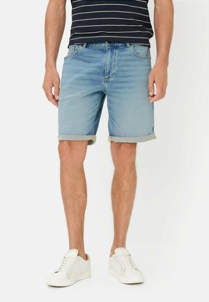 Man die lichtblauwe denimshorts met opgerolde pijpen, witte sneakers en een zwart-wit gestreept shirt draagt, staande tegen een witte achtergrond.