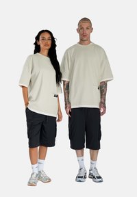 Svetlo béžové oversized tričká s kontrastnými manžetami, v kombinácii s čiernymi cargo šortkami. Výrazné tetovania a detaily vrecka na tričkách.