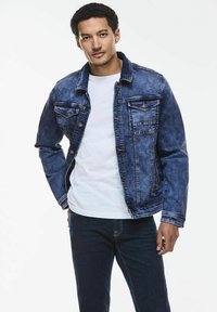 Jeansjacka i mörkblått med en blekt finish, två framfickor, knappstängning och en klassisk kragsdesign. Buren över en vit skjorta.