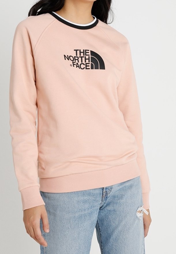 Personne portant un sweat-shirt The North Face rose clair avec des détails noirs et blancs au col et un jean bleu clair déchiré.