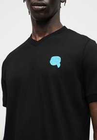 Μαύρο πουκάμισο με V λαιμόκοψη και μικρό μπλε κεντητό σχέδιο ενός κεφαλιού που φορά γυαλιά προστασίας και μαντήλι στο κεφάλι στο αριστερό στήθος, φορεμένο από άτομο με σκούρο δέρμα.