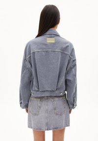 Chaqueta de mezclilla azul claro con un corte relajado, costuras detalladas, un gran panel trasero y una etiqueta en la parte superior de la espalda. Combinada con una falda a juego.