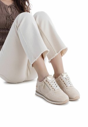Mujer sentada con pantalones cropped beige claro y zapatillas deportivas beige a juego, con cordones, cremallera lateral y patrones texturizados.