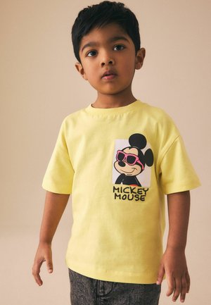 Young boy with dark hair wearing a yellow Mickey Mouse T-shirt with pink sunglasses graphic and dark pants, standing against a plain background.
