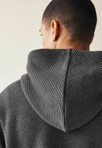 Grå tekstureret hoodie med et diamantmønster, en tætsiddende hætte og en søm ned i midten. Blødt stof synligt ved udskæringen.
