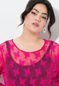 Durchsichtige rosa Bluse mit floralem Stickereimuster und Wellenkragen. Das Model trägt silberne Creolen vor einem weißen Hintergrund.