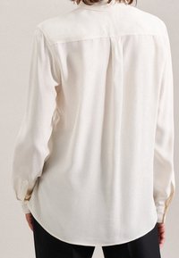 Person wearing a long-sleeve, cream-colored blouse with a back pleat and buttoned cuffs, paired with black pants, shown from behind.