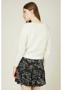 Pull en tricot crème avec des manches texturées, associé à une jupe noire à volants ornée d'un motif de feuilles blanches. Vue de dos.