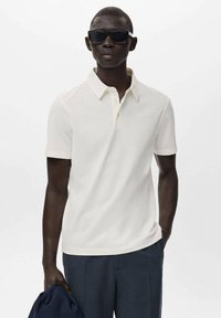 White polo shirt with a classic collar, short sleeves, and a smooth texture. Model wears black sunglasses and navy trousers, holding a dark bag.