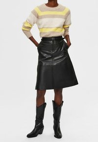 Gestreifter Strickpullover in Beige, Weiß und Gelb. Schwarzer Lederrock, knielang, mit einem Gürtel. Schwarze Cowboystiefel mit spitzer Zehe.