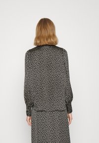Blusa negra con pequeño patrón floral crema, mangas largas, puños fruncidos y un dobladillo recto. Tela suave con una textura blanda.