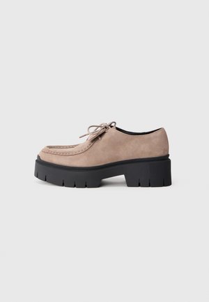 Chaussure beige en daim à lacets avec semelle épaisse noire et semelle extérieure crantée, montrée de profil sur un fond gris clair uni.