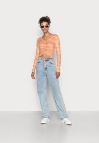 Top laranja de mangas longas com design franzido e detalhe de laço, combinado com jeans largos azul-claro e ténis brancos. Óculos de sol complementam o visual.