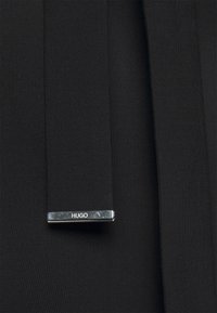 Mustat kangassiteet kiiltävällä hopeisella soljella, jossa on kaiverrus "Hugo", suunniteltu säädettävää istuvuutta ja turvallista kiinnitystä varten.