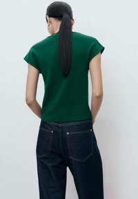 Green short-sleeve top with a round neckline, paired with dark denim jeans featuring back pockets and subtle stitching details.