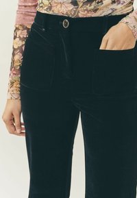 Osoba ubrana w czarne aksamitne spodnie z dużymi przednimi kieszeniami i kwiatową bluzkę z długim rękawem, ręka częściowo w prawej kieszeni.