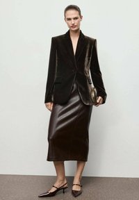 Mujer vistiendo un blazer de terciopelo marrón oscuro y una falda midi de cuero a juego, sosteniendo un pequeño clutch y llevando tacones de tipo slingback con punta.