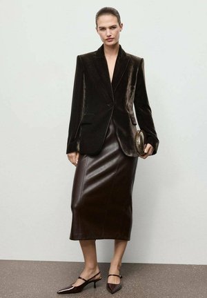 Femme portant un blazer en velours brun foncé et une jupe midi en cuir assortie, tenant une petite pochette, avec des escarpins à bout pointu et bride arrière.