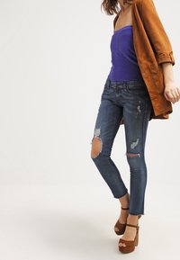 Jeans Skinny rasgados de color azul oscuro con detalles desgastados, combinados con una blusa sin tirantes de color púrpura y una chaqueta oversized de ante marrón. Tacones de plataforma marrones.