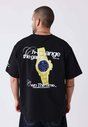Homme portant un t-shirt noir avec un grand motif de montre jaune et bleue et un texte blanc "Change the game, Own The Time" dans le dos.