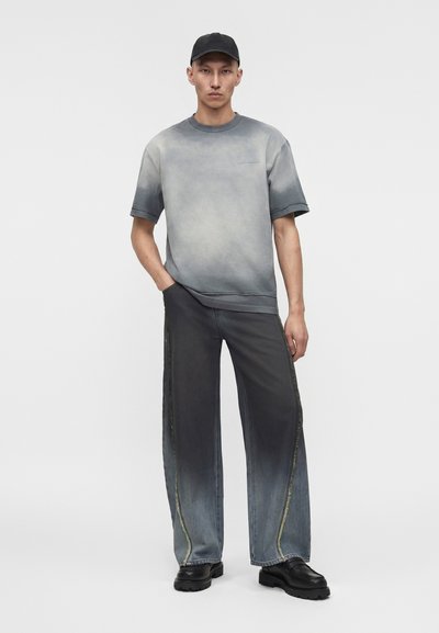 Άνδρας που φοράει ξεθωριασμένο μαύρο καπέλο, γκρι κοντομάνικη μπλούζα με διαβάθμιση, μαύρο πλυμένο τζιν με φαρδιά πόδια και ραφές στα πλάγια, και μαύρα χοντρά παπούτσια, όρθιος.