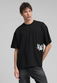 T-shirt in cotone oversize nero con una stampa grafica bianca nell'angolo in basso a sinistra. Scollatura rotonda semplice e maniche corte.