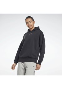 Schwarzer Kapuzenpullover mit geripptem Saum und Bündchen, ausgestattet mit einem kleinen weißen Logo auf der Brust, kombiniert mit grauen Sweatpants.