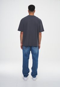 En man står vänd bort från kameran och bär en mörkgrå oversized T-shirt, blå jeans och vita sneakers framför en enkel vit bakgrund.