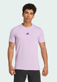 Camiseta deportiva de color púrpura claro, hecha de una tela suave, con un cuello redondo y mangas cortas. Presenta un pequeño logo negro de Adidas en el pecho.