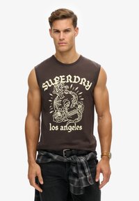 Camiseta sin mangas marrón con un estampado gráfico blanco de un dragón y rosas, con el texto "SUPERDRY los ángeles", combinada con jeans oscuros y una camisa de cuadros atada a la cintura.
