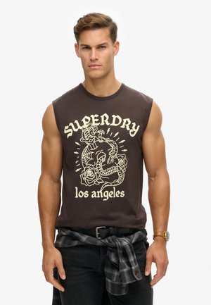 Brun ærmeløs t-shirt med hvid tryk af en drage og roser, med teksten "SUPERDRY los angeles", båret med mørke jeans og et ternet tørklæde bundet om livet.
