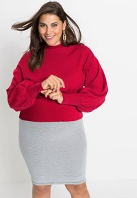 Maglione rosso a maglia con maniche a sbuffo, abbinato a una gonna in rete grigia attillata. Gli orecchini presentano un design geometrico. Sfondo neutro.