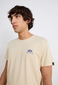 Beige pamut póló kerek nyakkal, a bal mellkason kék "Alpha Industries" logóval és részlettel. Rövid ujjú.