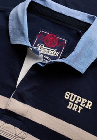 Maillot de rugby bleu marine avec un col bleu clair, des rayures horizontales beige et "SUPER DRY" brodé sur la poitrine.