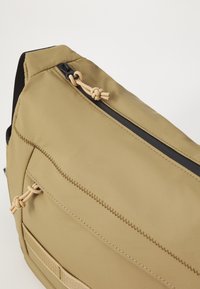 Khaki sling bag with textured fabric, black zipper, and tan cord accents. Features a front pocket with visible stitching and a patch.