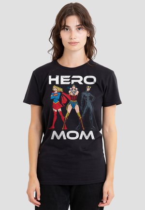 Woman wearing a black T-shirt with "HERO MOM" text and illustrations of Supergirl, Wonder Woman, and Catwoman standing in strong poses.