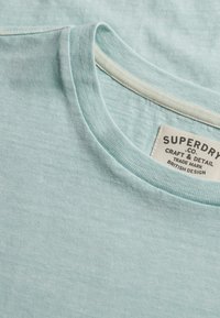 T-shirt bleu clair col rond avec une étiquette de marque Superdry visible cousue à l'intérieur du col.