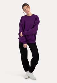 Sudadera oversize viola con polsini e orlo a coste, abbinata a pantaloni joggers neri e sneakers bianche, in piedi su uno sfondo neutro.