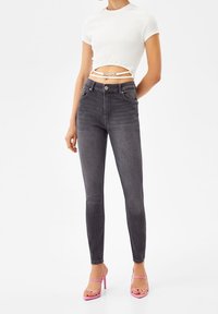 Jeans skinny de tiro alto negros con una textura suave, combinados con una blusa corta blanca ajustada que presenta detalles recortados y tirantes finos. Sandalias de tacón alto rosas.