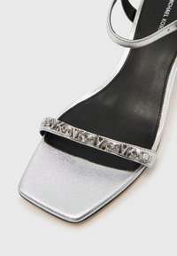 Ασημένιο σανδάλι με ψηλό τακούνι και μεταλλική επίστρωση, με τετράγωνη μύτη και μαύρο εσωτερικό. Διαθέτει διακοσμητικό λουράκι με ανάγλυφο λογότυπο "MK".