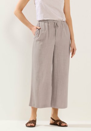 Kobieta ubrana w jasnoszare, szerokie spodnie o długości do połowy łydki z wiązanym paskiem w talii i brązowe płaskie sandały, stojąca na jednolitym tle.