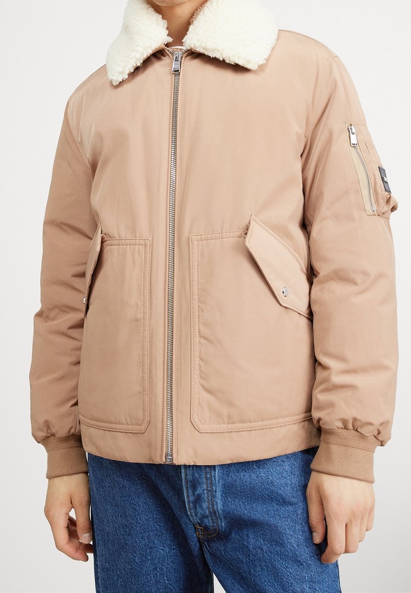 Beige bomberjacka med krage av vit fårskinn, dragkedja framtill, två stora framfickor och ribbade muddar. Slät textur, lätt stoppning.