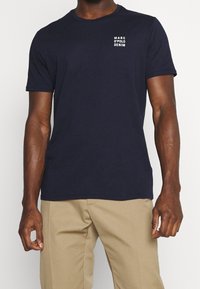 Navy sininen puuvillapaita lyhythihaisilla, pyöreällä kaula-aukolla ja pieni valkoinen logo vasemmassa rinnassa. Sileä kangas, normaali malli.