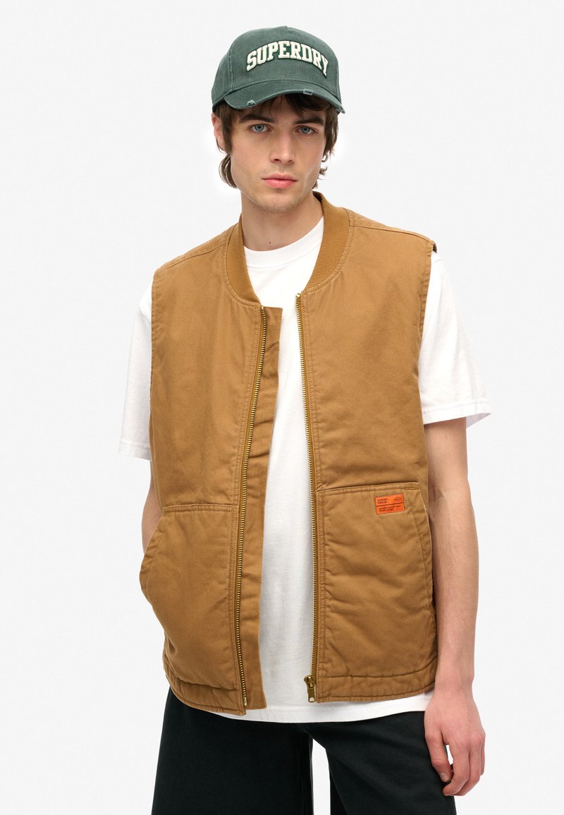 Chaleco marrón sin mangas con cremallera frontal, bolsillos laterales y una etiqueta naranja. Lleva puesto sobre una camiseta blanca, combinado con una gorra verde.