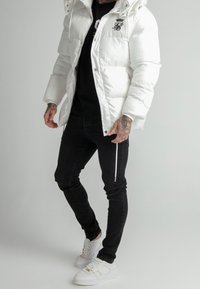 Chaqueta acolchada blanca con capucha, que presenta un cierre de cremallera y un logo en el pecho. Combinada con jeans negros ajustados y zapatillas blancas.