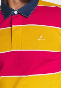 Randig polo t-shirt med rosa, gula och vita ränder; denimkrage; GANT-logotyp broderad i vitt på bröstet; blandmaterial av bomull.